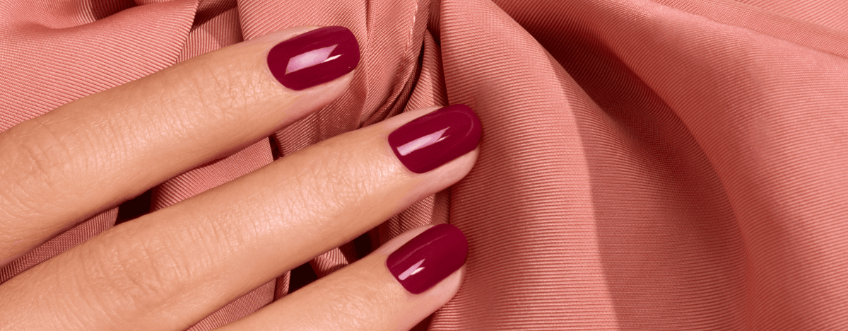A hand with glossy, deep red manicured fingernails resting against draped, muted pink fabric.
