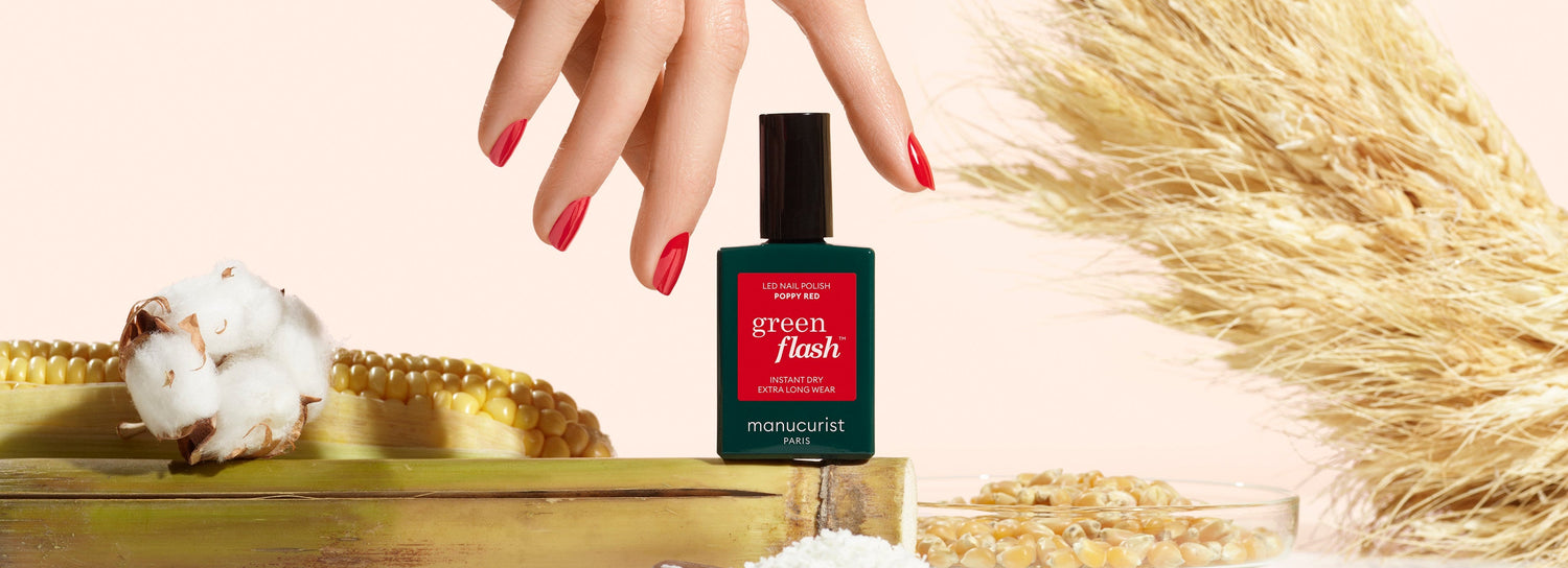 A hand with red-painted nails hovers above a green nail polish bottle labeled green flash. Surrounding it are a green K4VN egg, dried wheat, cotton, corn, and grains on a light background.