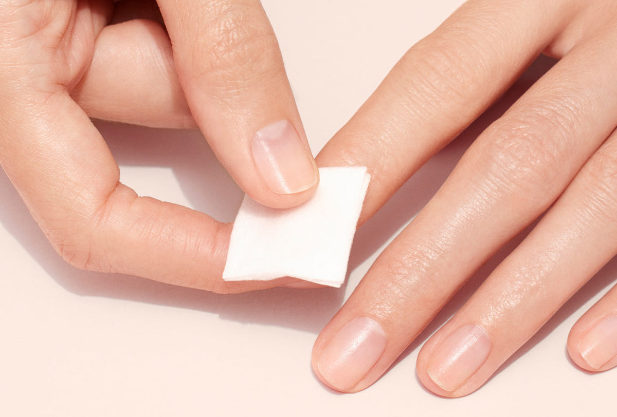 A hand uses a small white cotton pad to clean or remove nail polish from fingernails on the other hand, against a light beige background.