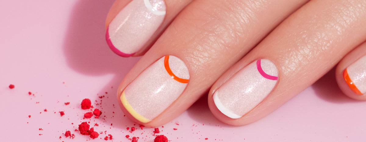 A close-up of a hand with light pink, sparkly nail polish and colorful, minimalist French tips in yellow, orange, pink, and white. Red powder is scattered on the pink surface.
