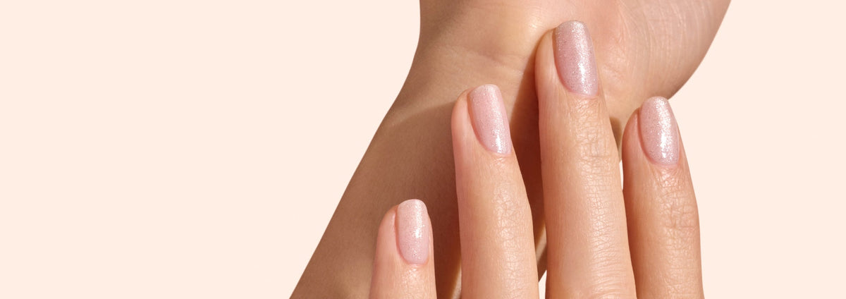 A close-up of a hand with neatly manicured nails painted in a light, shimmery nude color against a soft beige background.