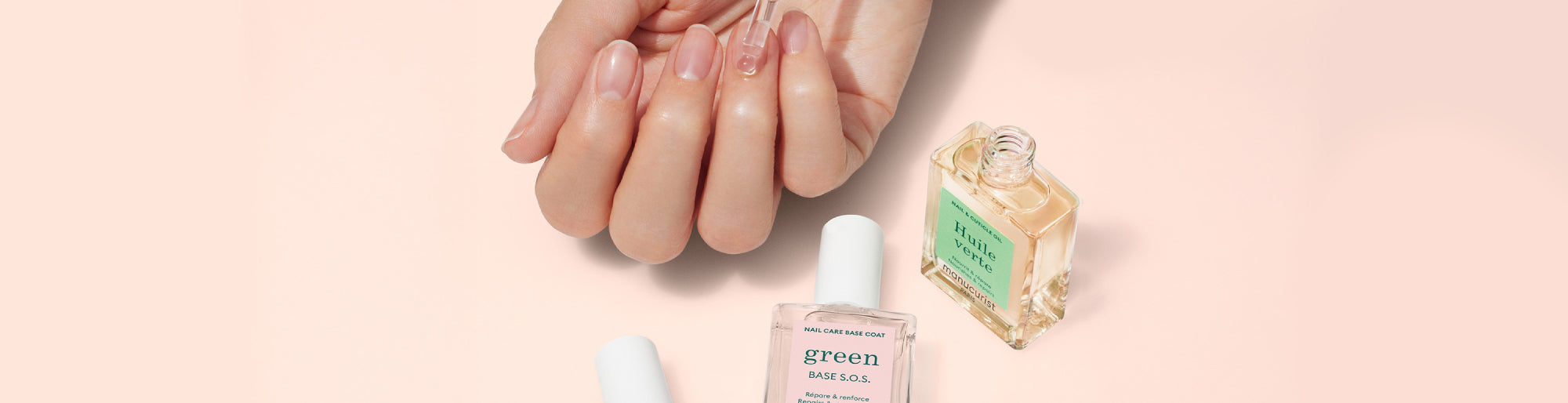 A hand with neatly manicured nails rests on a light pink surface next to two bottles of clear and light yellow nail polish with white caps.