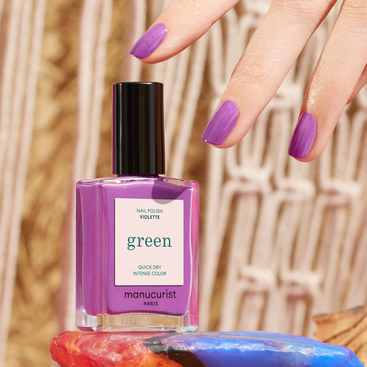A hand with retro purple painted nails hovers above a Violette nail polish bottle by Manucurist, set on a colorful surface with a woven background.