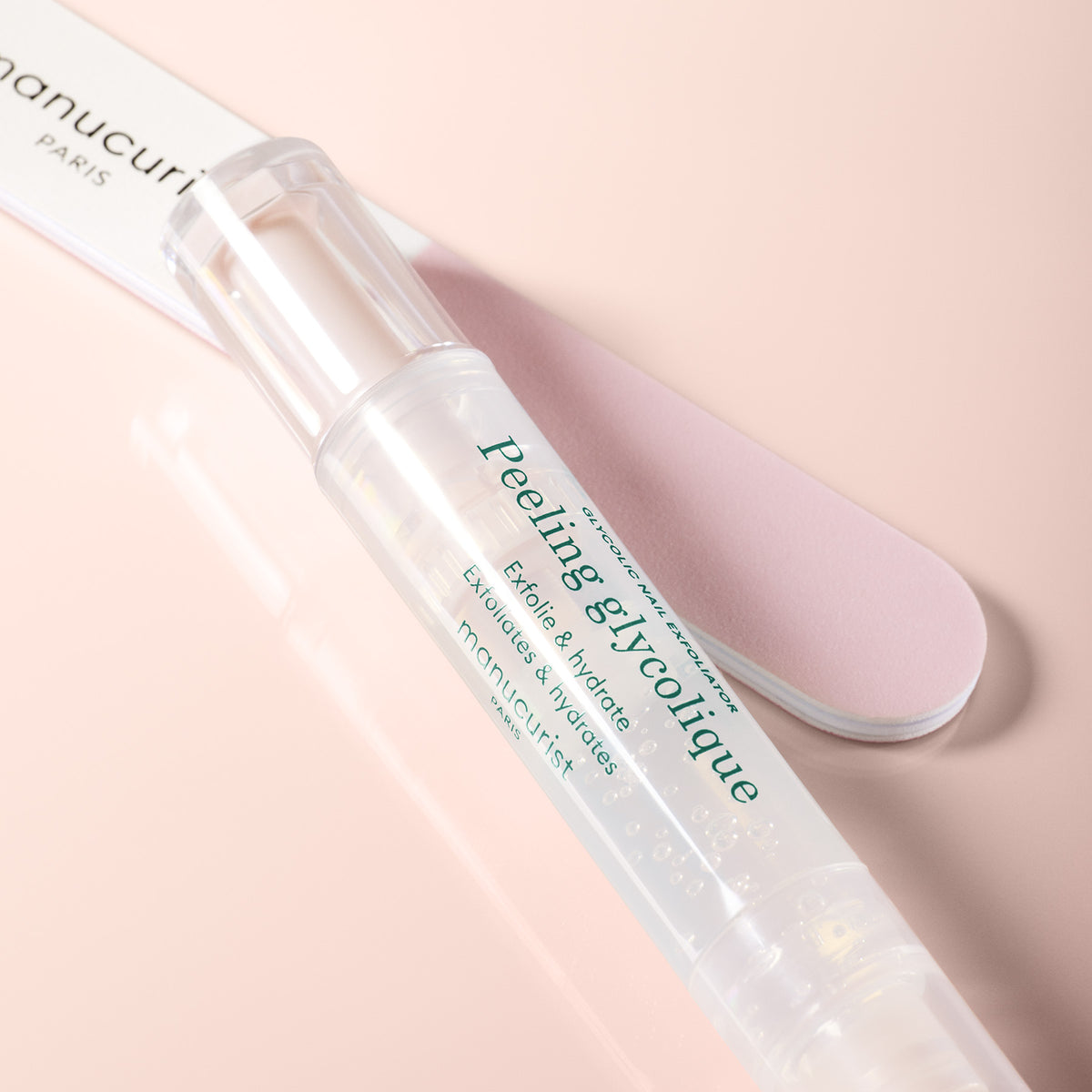 Un Prep Glow Routine transparent en forme de stylo de Manucurist repose sur une surface rose à côté d’une lime à ongles rose clair — idéal pour exfolier et hydrater les ongles et les cuticules afin d’obtenir des ongles brillants dans votre routine de soin.