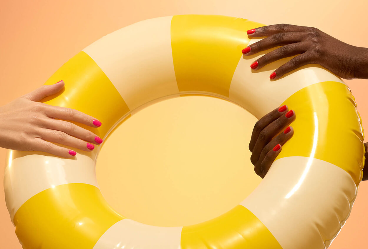 Two hands with brightly painted nails, one light-skinned and one dark-skinned, hold opposite sides of a yellow and white inflatable pool ring against a warm, gradient background.