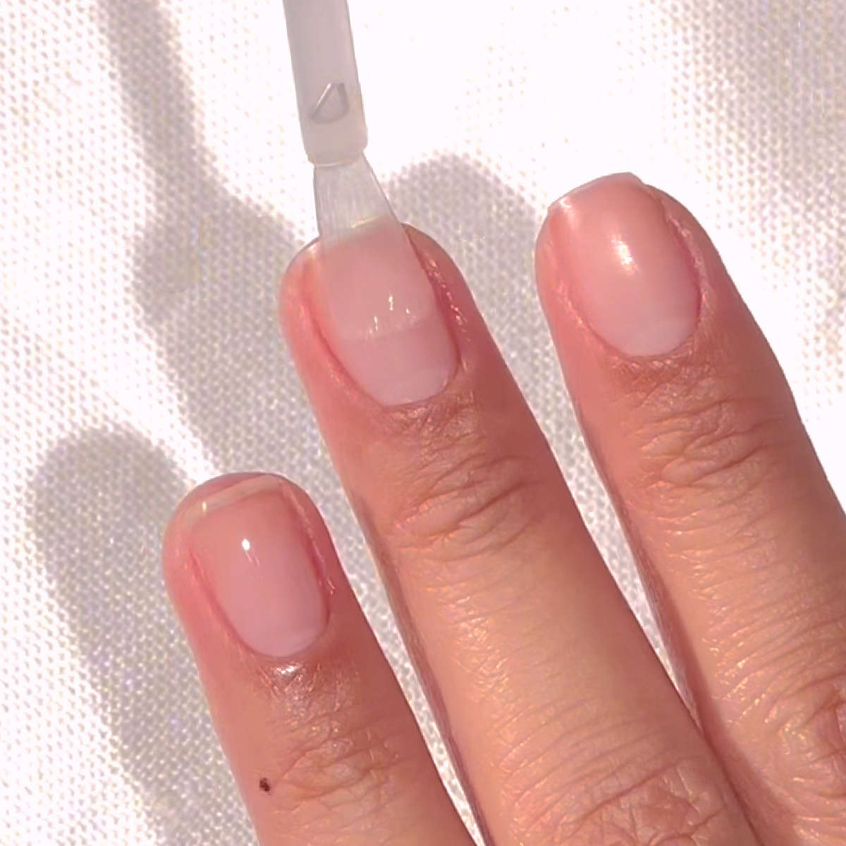 A hand with neatly manicured nails is shown as a clear nail polish is being applied to the middle fingernail using a brush. The background is a white textured surface.