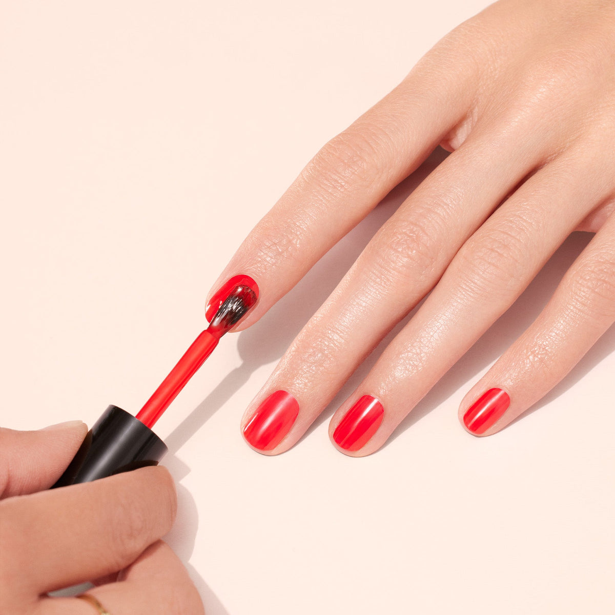 Les ongles d’une main sont peints avec un vernis rouge vif. Une autre main tient le pinceau et applique le vernis sur les ongles, sur un fond clair et neutre.