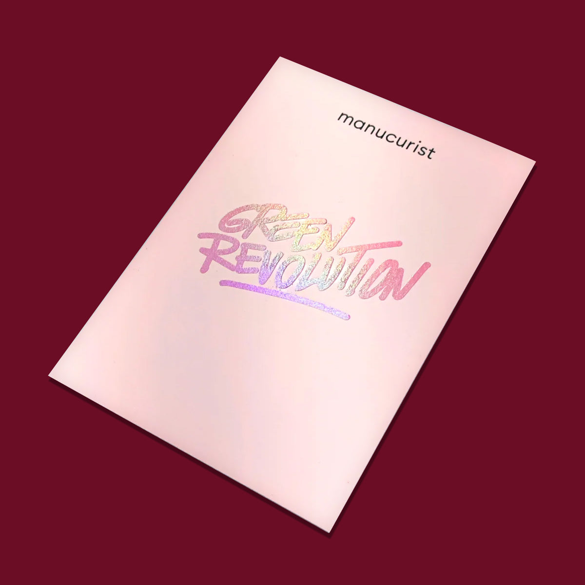 Un livret rose pâle sur fond rouge foncé met en avant la carte cadeau Manucurist US, avec le texte holographique "GREEN REVOLUTION" — un choix inspirant pour offrir plus responsable.