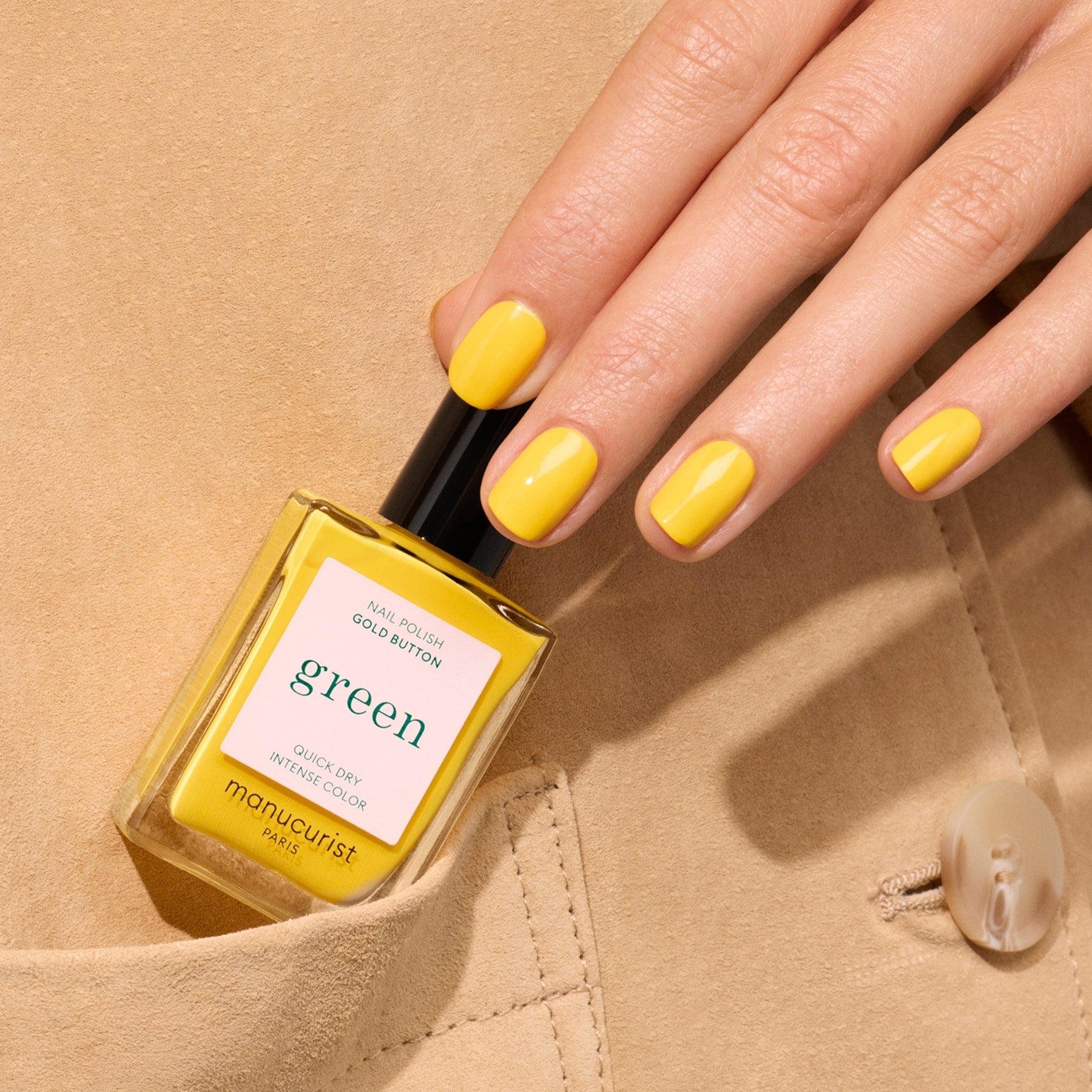 A hand with neatly manicured nails painted in Gold Button by Manucurist holds a matching yellow Gold Button nail polish bottle, resting against a beige fabric background.