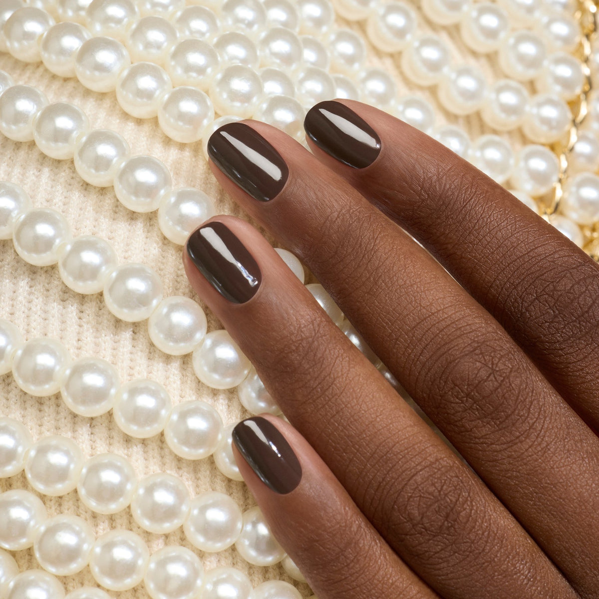 A hand with Manucurist’s Mocha gel polish rests on a white knit fabric adorned with rows of white pearls.
