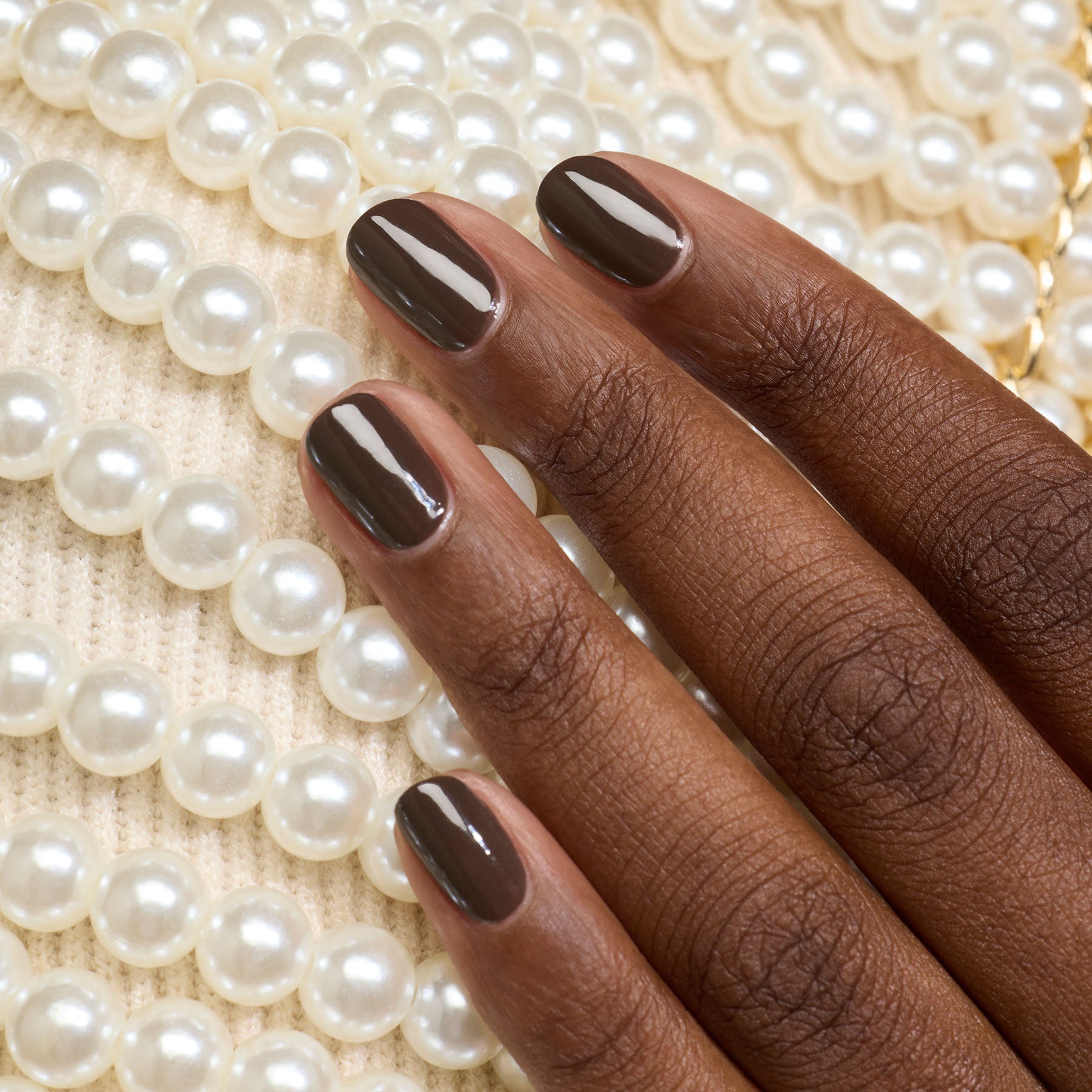 A hand with Manucurist’s Mocha gel polish rests on a white knit fabric adorned with rows of white pearls.