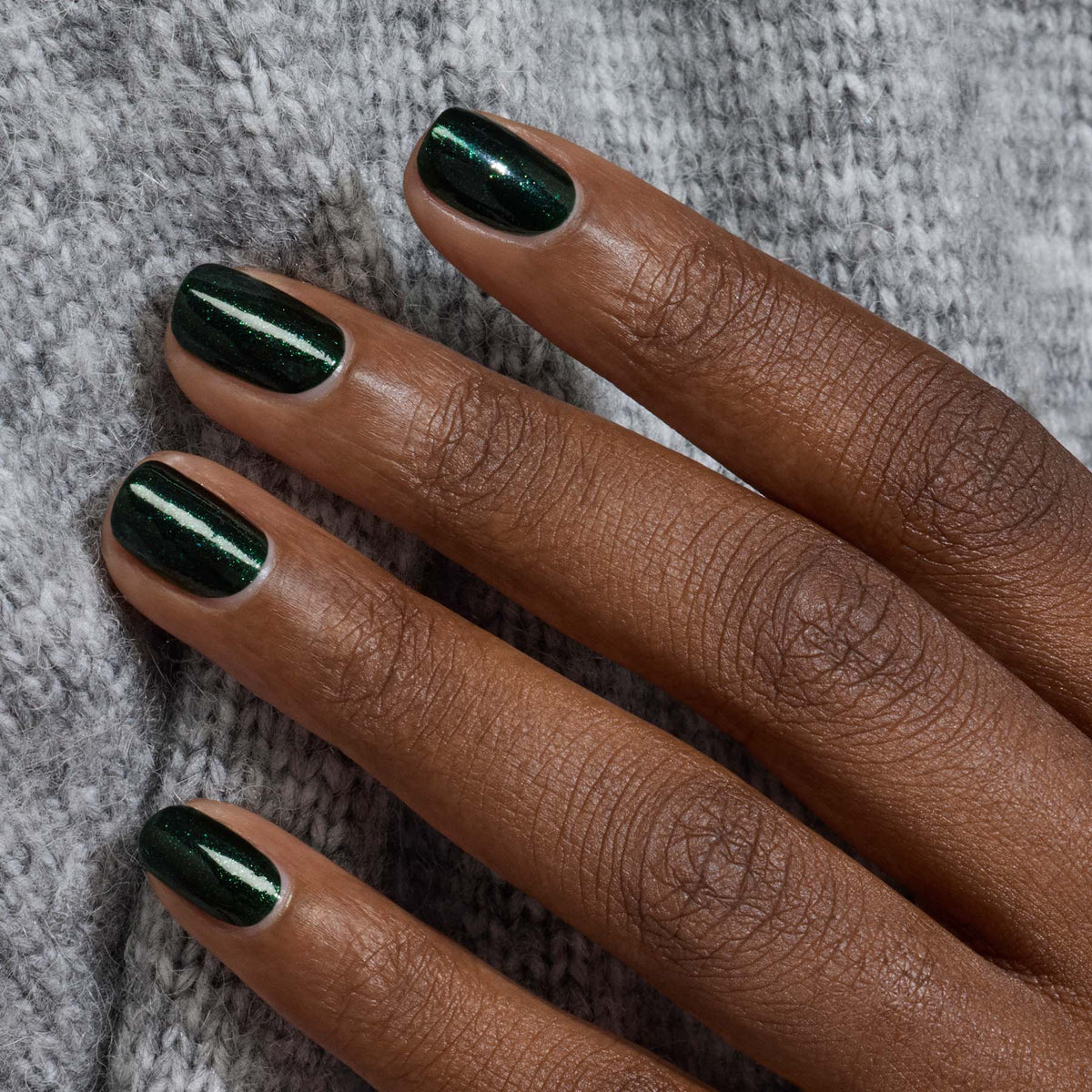 Gros plan d’une main avec le vernis gel Manucurist Poison brillant vert forêt, posée sur un tissu en maille gris clair.