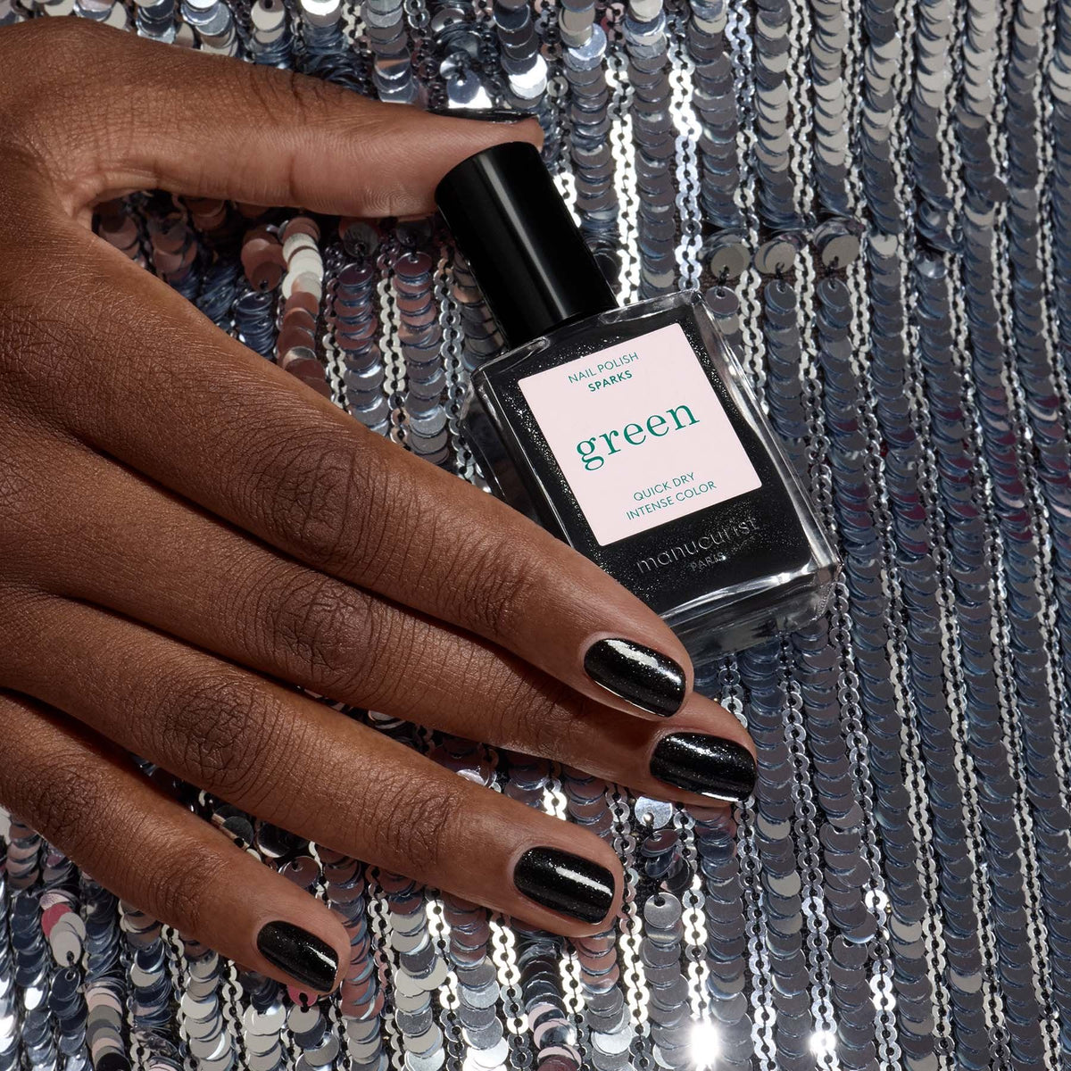 A hand with glossy black nail polish rests on silver sequins, holding a bottle of Manucurist’s Sparks nail polish that glimmers with a shimmering finish.