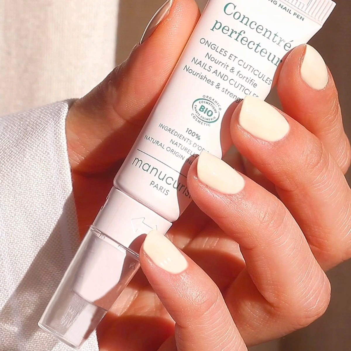 A person with neatly manicured, pale nude nails holds a tube of Manucurist Paris nail and cuticle cream, gently squeezing some product onto their other hand.
