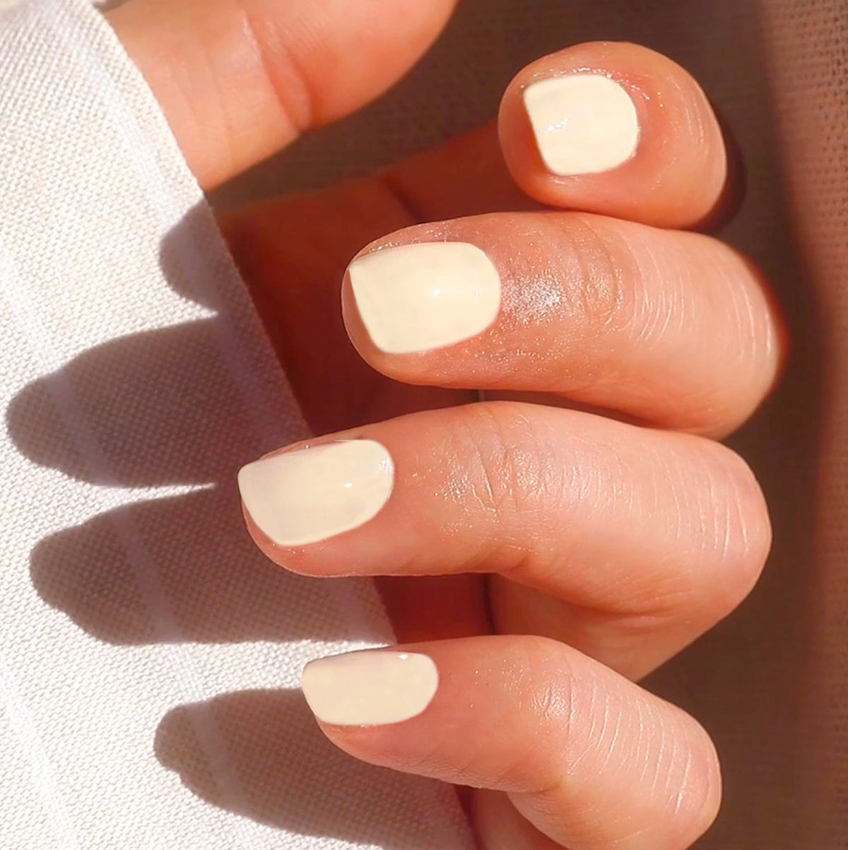 A close-up of a hand with short, neatly filed nails painted in a glossy, pale cream color, holding a piece of white fabric.