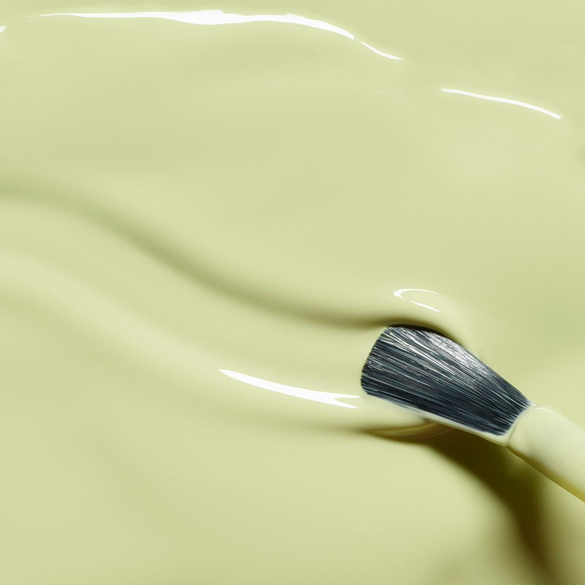 A makeup brush rests on a smooth, creamy Pistachio surface with yellow undertones from Manucurist, featuring gentle waves and glossy reflections.