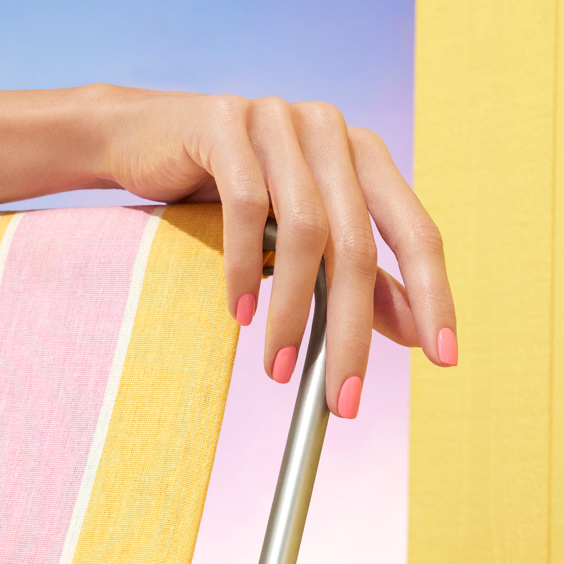A hand with short, neatly manicured nails painted in Manucurist’s Pop electric pink polish rests on a metal chair with yellow and pink striped fabric, set against a pastel sky backdrop.