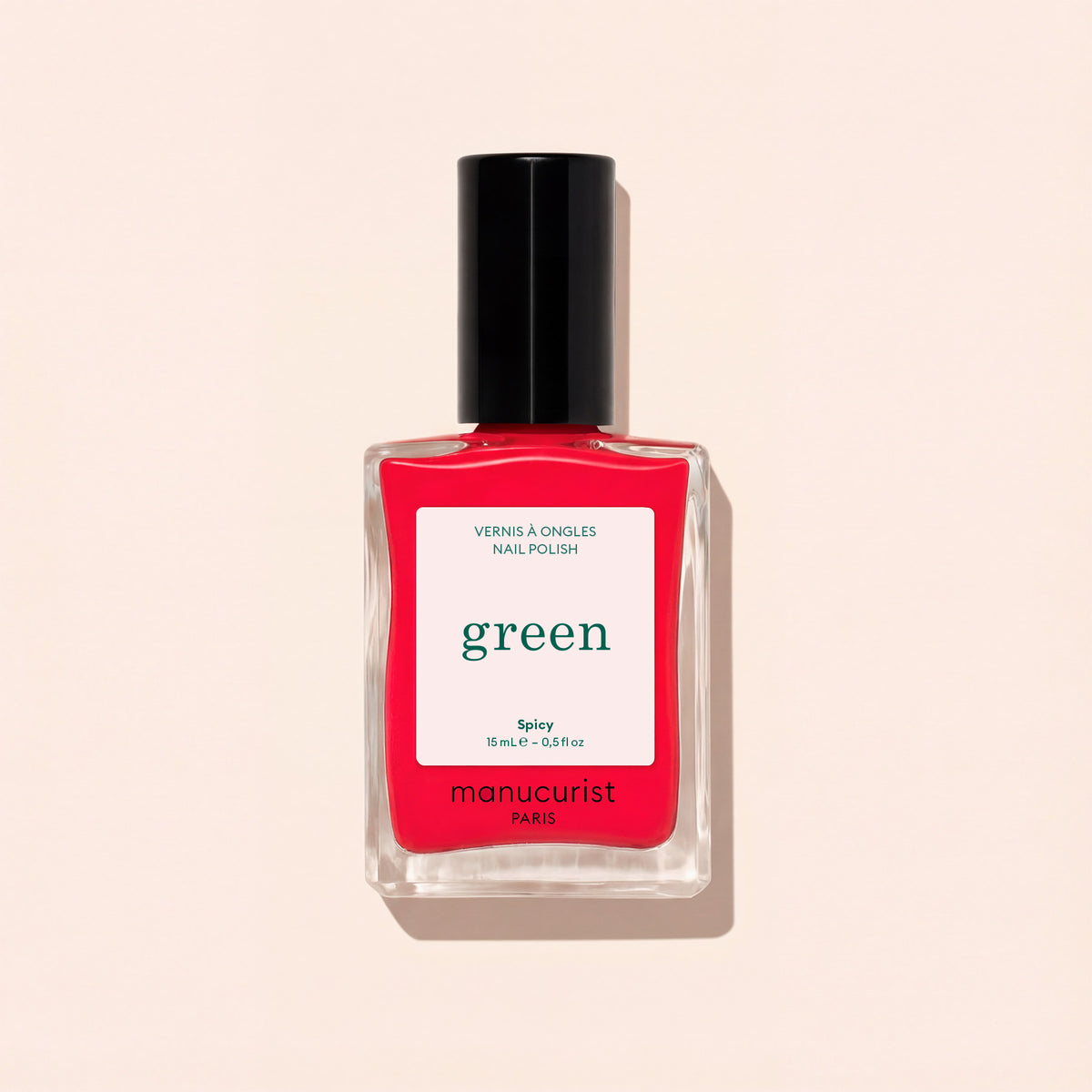 Un flacon de vernis à ongles Spicy de Manucurist, une teinte rouge pop aux sous-tons roses de la gamme Green™, avec un bouchon noir et une étiquette blanche, sur un fond beige clair.