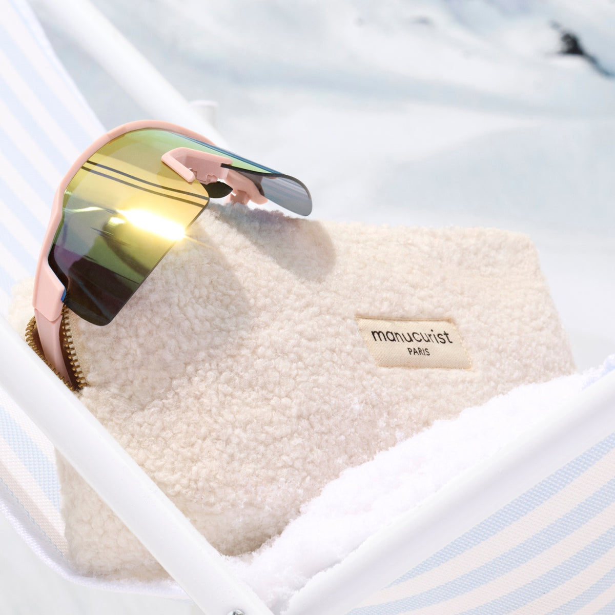 Une paire de lunettes de soleil réfléchissantes repose sur le doux Teddy Bag de Manucurist-us, posé sur une surface rayée blanche et bleue avec un décor enneigé — un accessoire d’hiver chic.