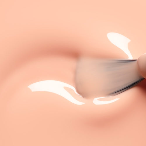 A close-up of a brush applying a glossy, clear liquid onto a smooth Milky Peach gel polish surface by Manucurist.