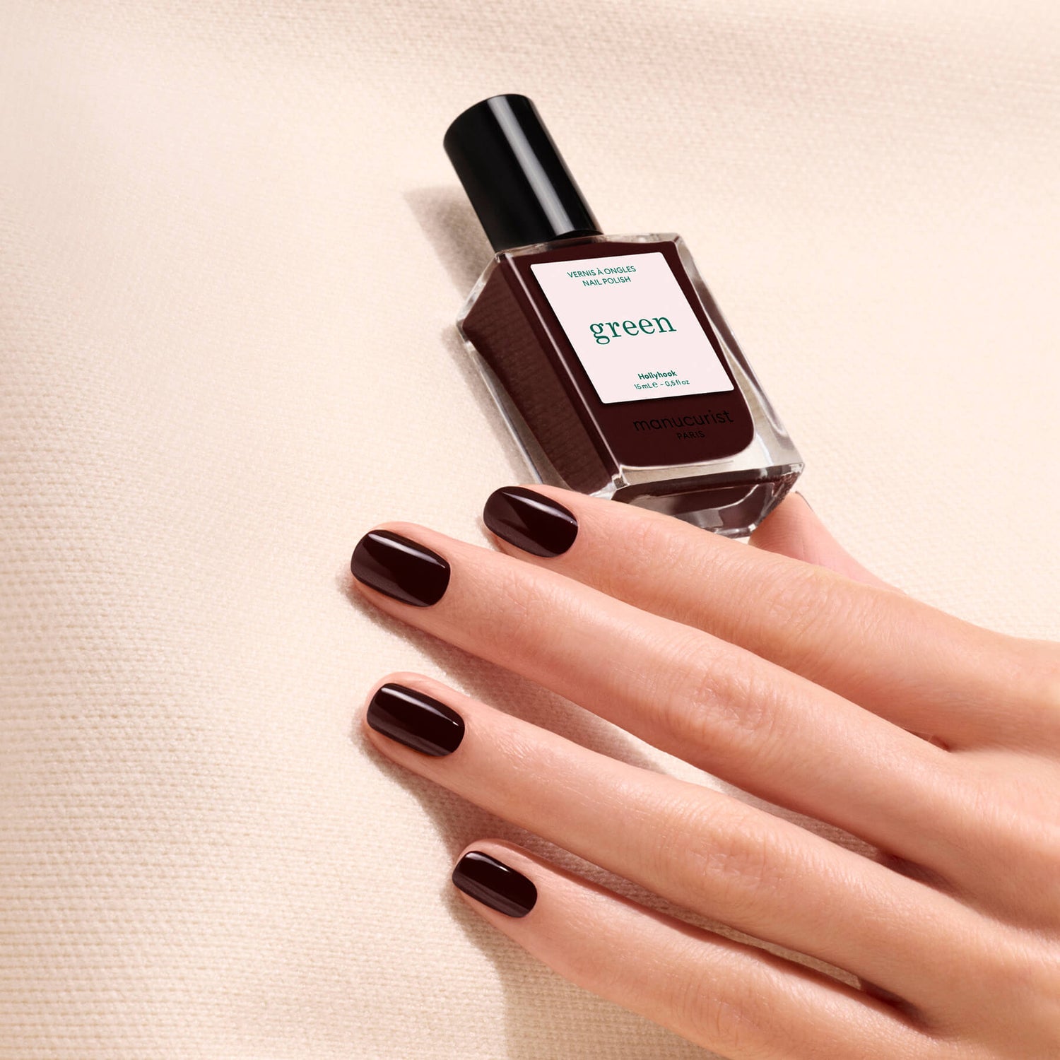 A hand with short, glossy dark brown nails rests on a textured beige surface next to a bottle of Manucurist Green™ nail polish in the shade “Hollyhock.”.