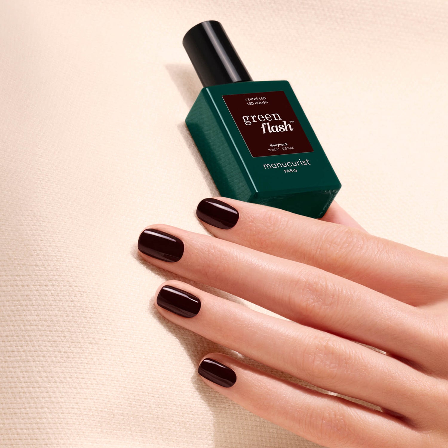 A hand with short, dark burgundy polished nails—reminiscent of a deep red nail polish—rests on a textured cream surface next to a green bottle of Manucurist’s Green Flash gel polish in the shade “Hollyhock.”.