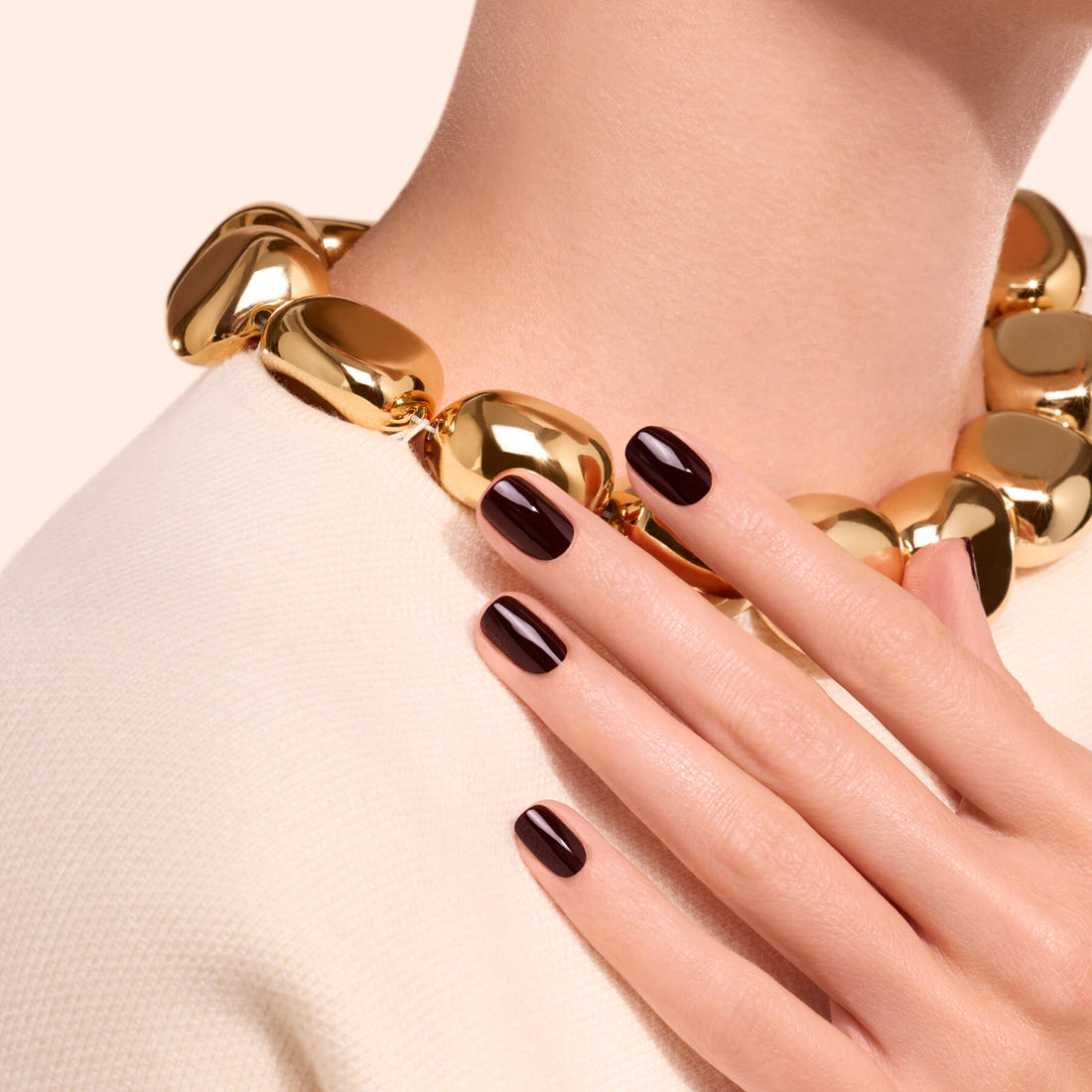 A hand adorned with Manucurist’s Hollyhock—a deep red nail polish—rests on a textured white top. The person wears a chunky gold chain necklace, set against a pale, simple background.
