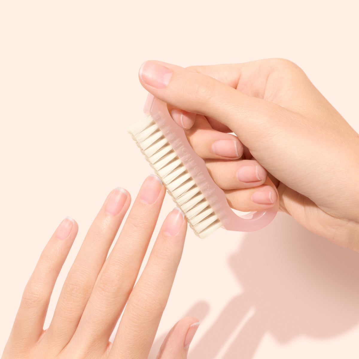 A person uses the Manucurist-us Nail Brush, featuring white vegan bristles, to clean their fingernails against a pale background. The hands and the brush are in focus, highlighting a simple manicure routine.