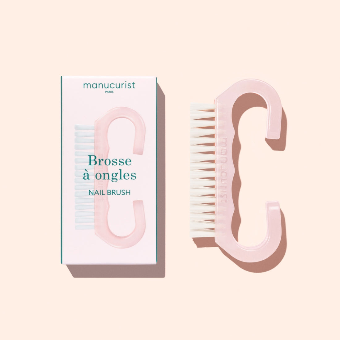 A translucent pink Nail Brush with vegan bristles and white tips from Manucurist-us rests next to its box, which displays an image of the brush and the words “Manucurist Paris, Brosse à ongles, NAIL BRUSH,” set against a pale background—ideal for enhancing your manicure routine.