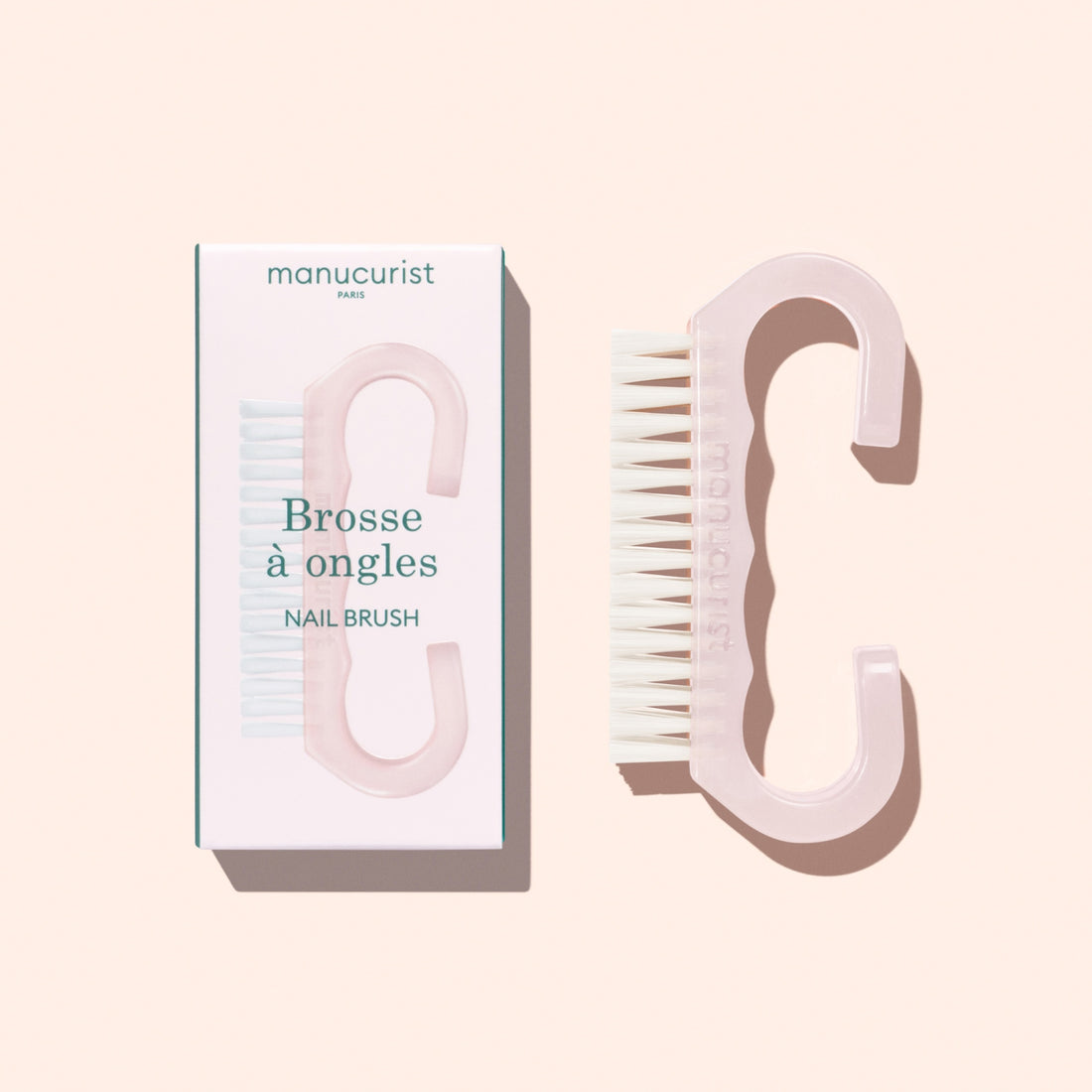 A translucent pink Nail Brush with vegan bristles and white tips from Manucurist-us rests next to its box, which displays an image of the brush and the words “Manucurist Paris, Brosse à ongles, NAIL BRUSH,” set against a pale background—ideal for enhancing your manicure routine.
