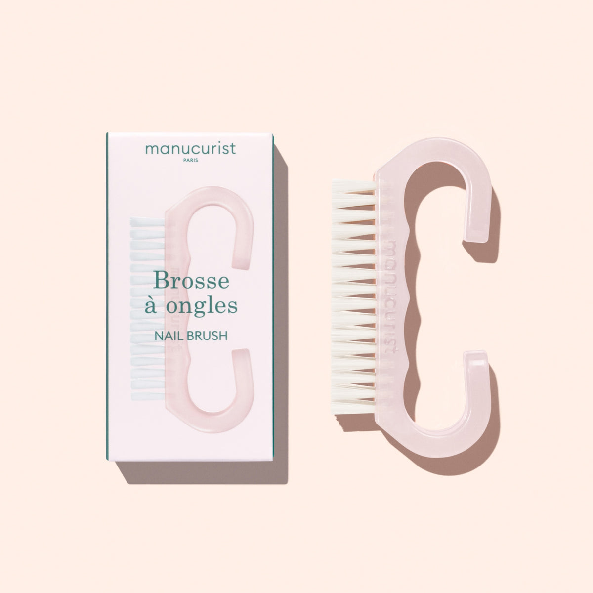 A translucent pink Nail Brush with vegan bristles and white tips from Manucurist-us rests next to its box, which displays an image of the brush and the words “Manucurist Paris, Brosse à ongles, NAIL BRUSH,” set against a pale background—ideal for enhancing your manicure routine.