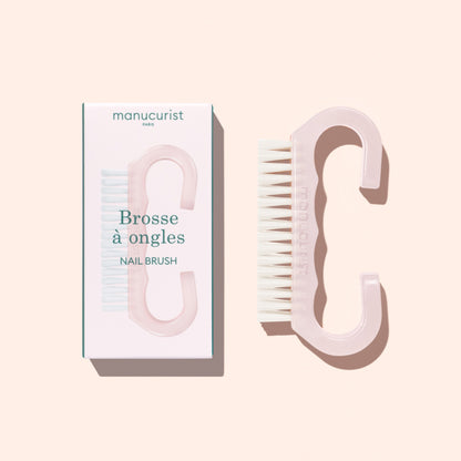 A translucent pink Nail Brush with vegan bristles and white tips from Manucurist-us rests next to its box, which displays an image of the brush and the words “Manucurist Paris, Brosse à ongles, NAIL BRUSH,” set against a pale background—ideal for enhancing your manicure routine.