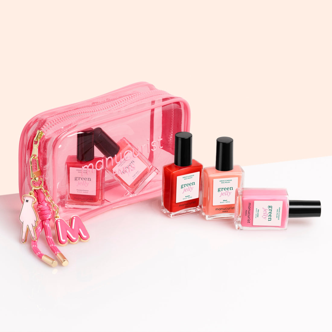 Three Manucurist Jelly Charm nail polishes in red, pink, and clear are arranged in front of a clear pink Manucurist manicure bag with a Jelly Charm keychain. The bag contains more nail polish bottles.