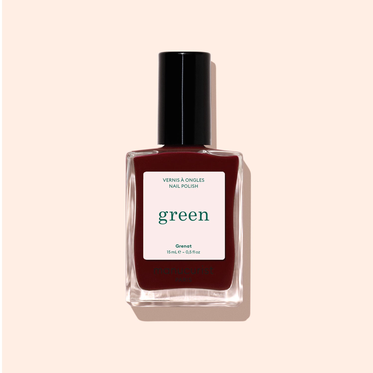 A bottle of Manucurist Grenat nail polish from the Green range, with a black cap, is displayed against a light beige background. The label reads “green” and “Nail Polish,” highlighting this earthy maroon red shade.