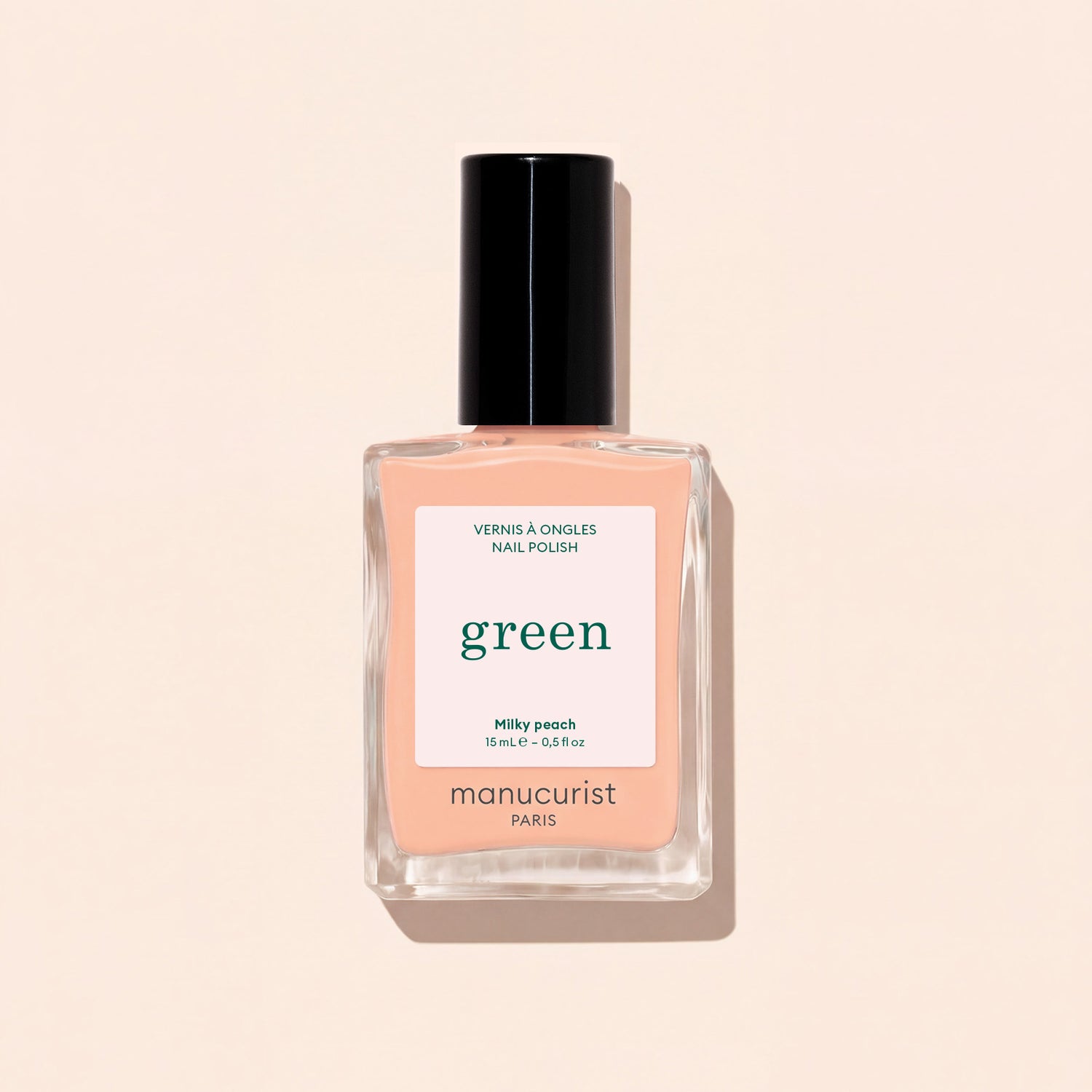 A bottle of Manucurist Milky Peach nail polish rests on a light beige surface. The clear bottle, featuring a black cap and a white label with green and peach lettering, beautifully highlights this soft peach shade from Manucurist.