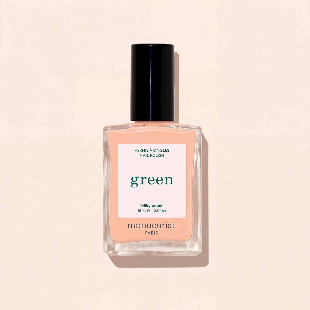 A bottle of Manucurist Milky Peach nail polish rests on a light beige surface. The clear bottle, featuring a black cap and a white label with green and peach lettering, beautifully highlights this soft peach shade from Manucurist.