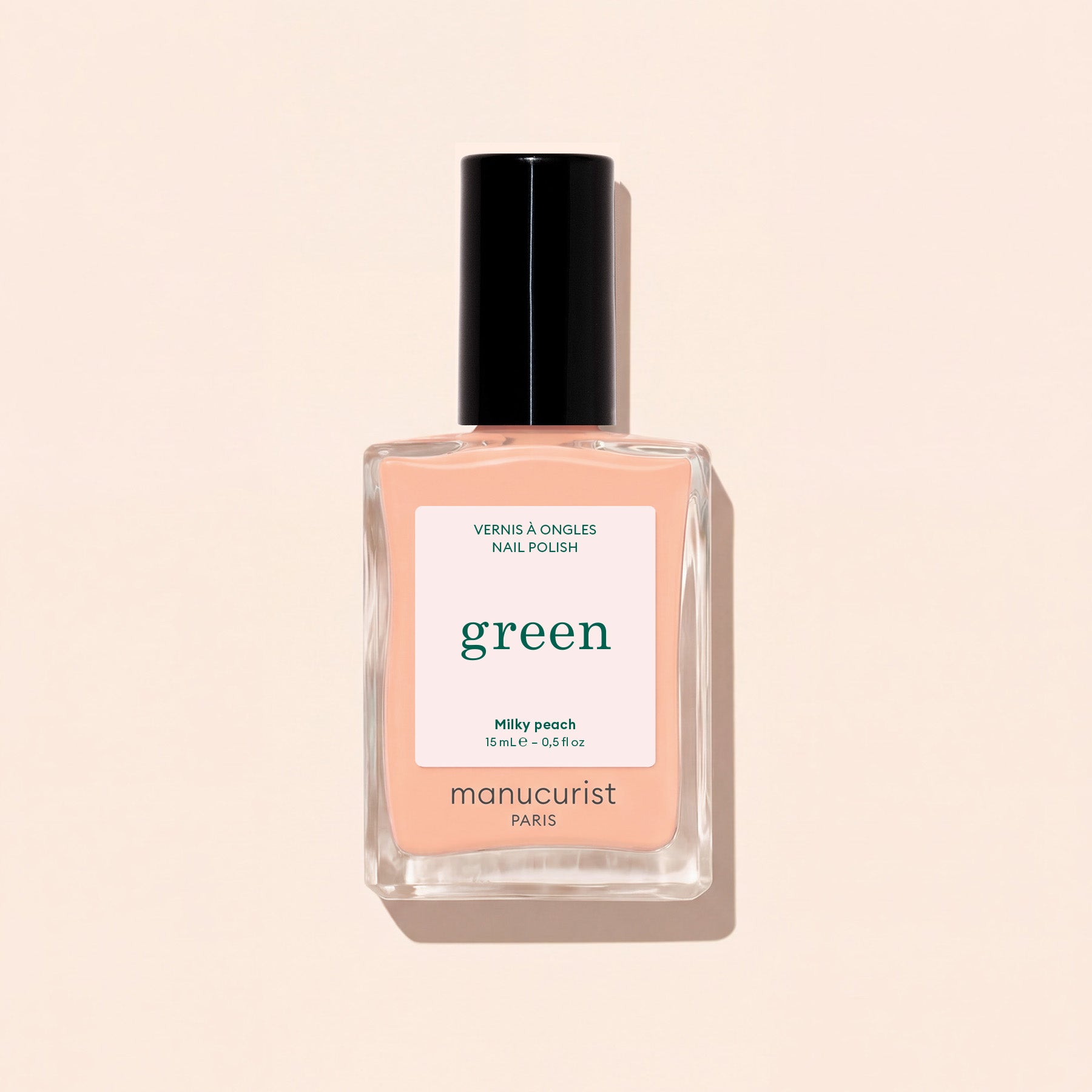 A bottle of Manucurist Milky Peach nail polish rests on a light beige surface. The clear bottle, featuring a black cap and a white label with green and peach lettering, beautifully highlights this soft peach shade from Manucurist.