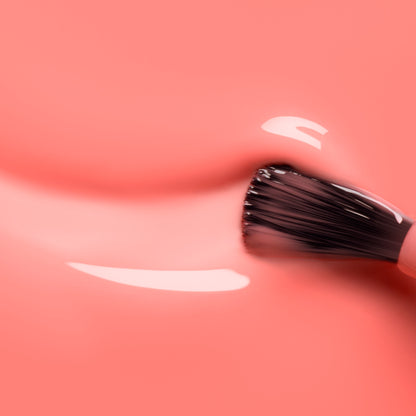 A close-up of a brush applying Manucurist Glacé, an ultra-glossy coral pink nail polish, with smooth, shiny strokes visible on the surface.
