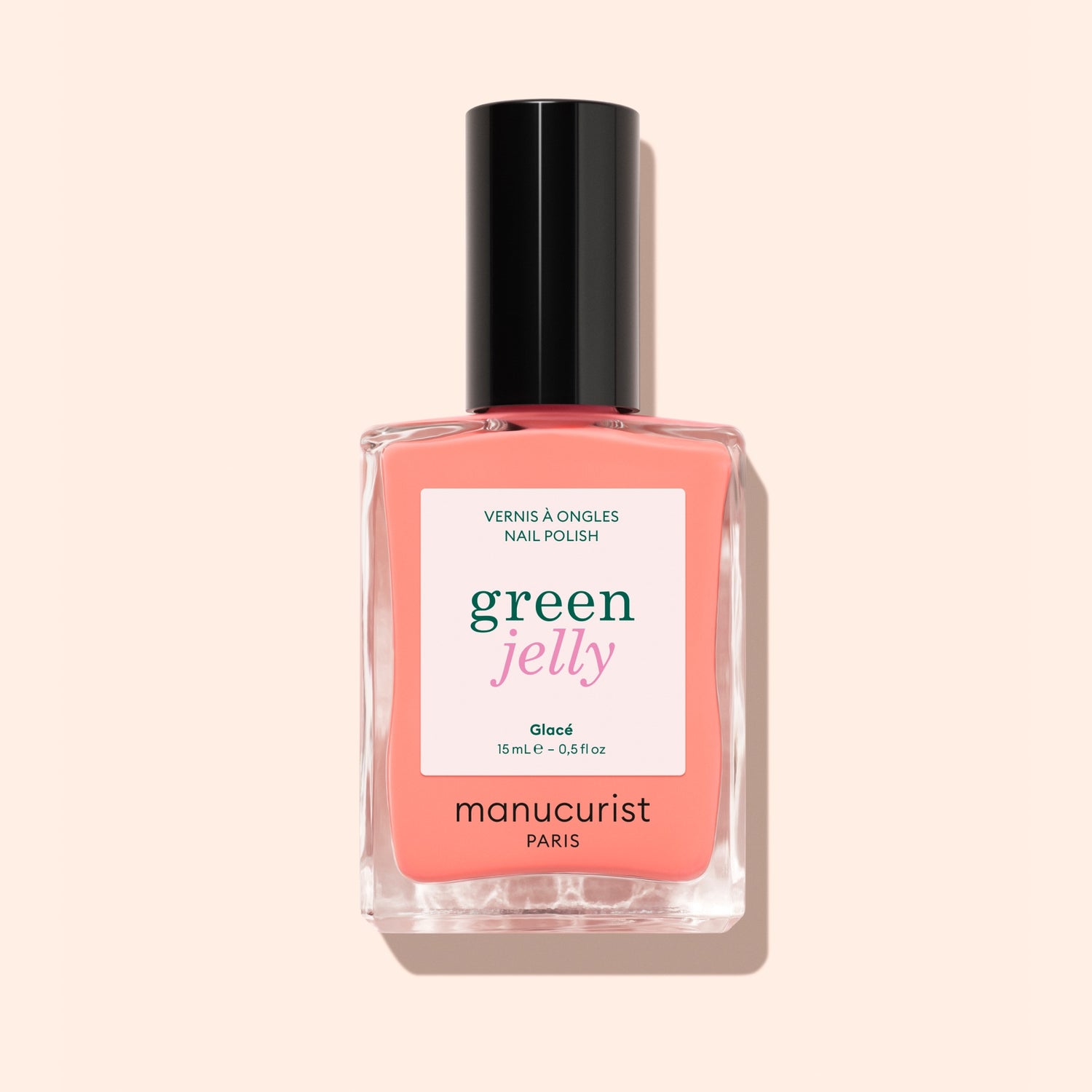 A bottle of Manucurist Glacé ultra-glossy nail polish in a pink-coral shade, featuring a black cap, is photographed against a pale peach background.