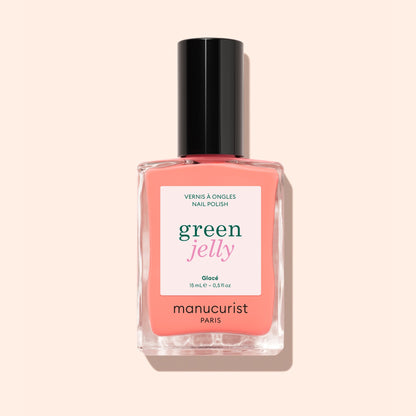A bottle of Manucurist Glacé ultra-glossy nail polish in a pink-coral shade, featuring a black cap, is photographed against a pale peach background.