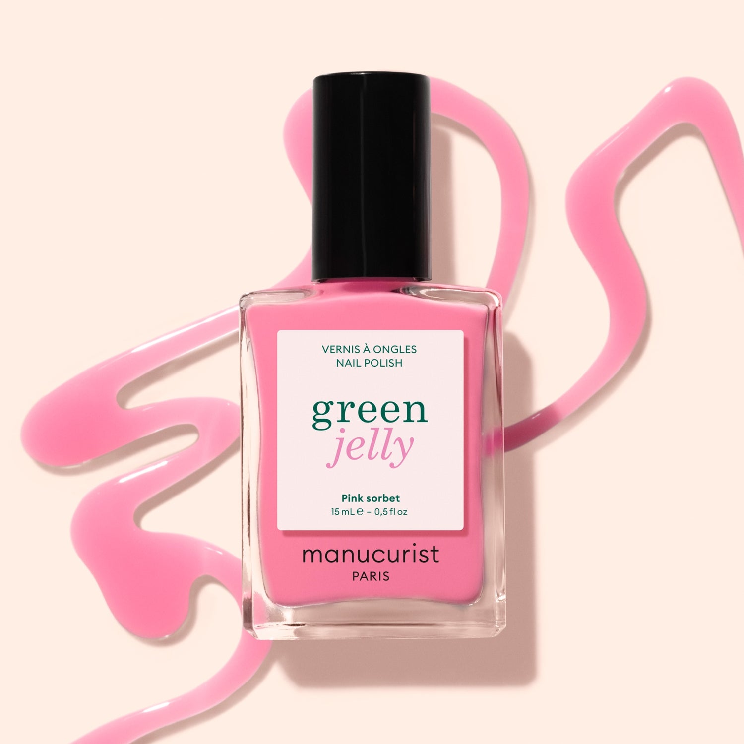 A bottle of Manucurist Pink Sorbet Jelly nail polish rests on a light beige surface, surrounded by translucent pink polish artistically spilled around it.