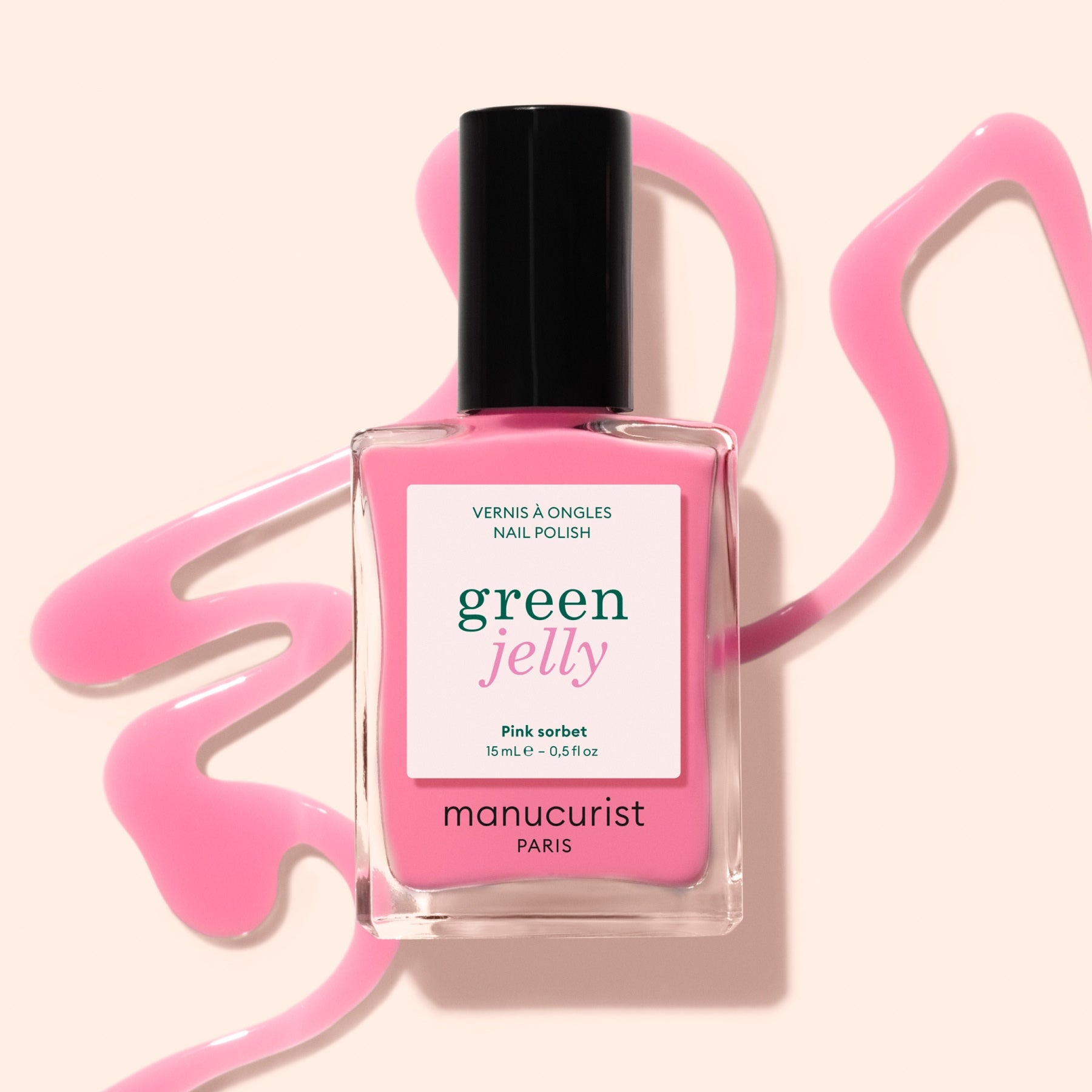 A bottle of Manucurist Pink Sorbet Jelly nail polish rests on a light beige surface, surrounded by translucent pink polish artistically spilled around it.