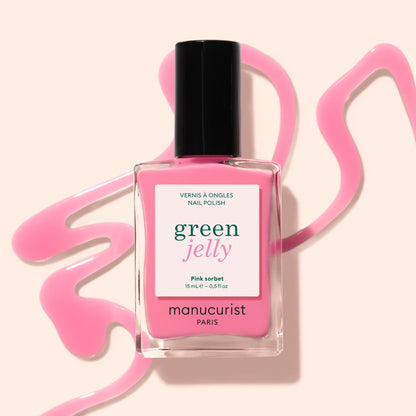 A bottle of Manucurist Pink Sorbet Jelly nail polish rests on a light beige surface, surrounded by translucent pink polish artistically spilled around it.