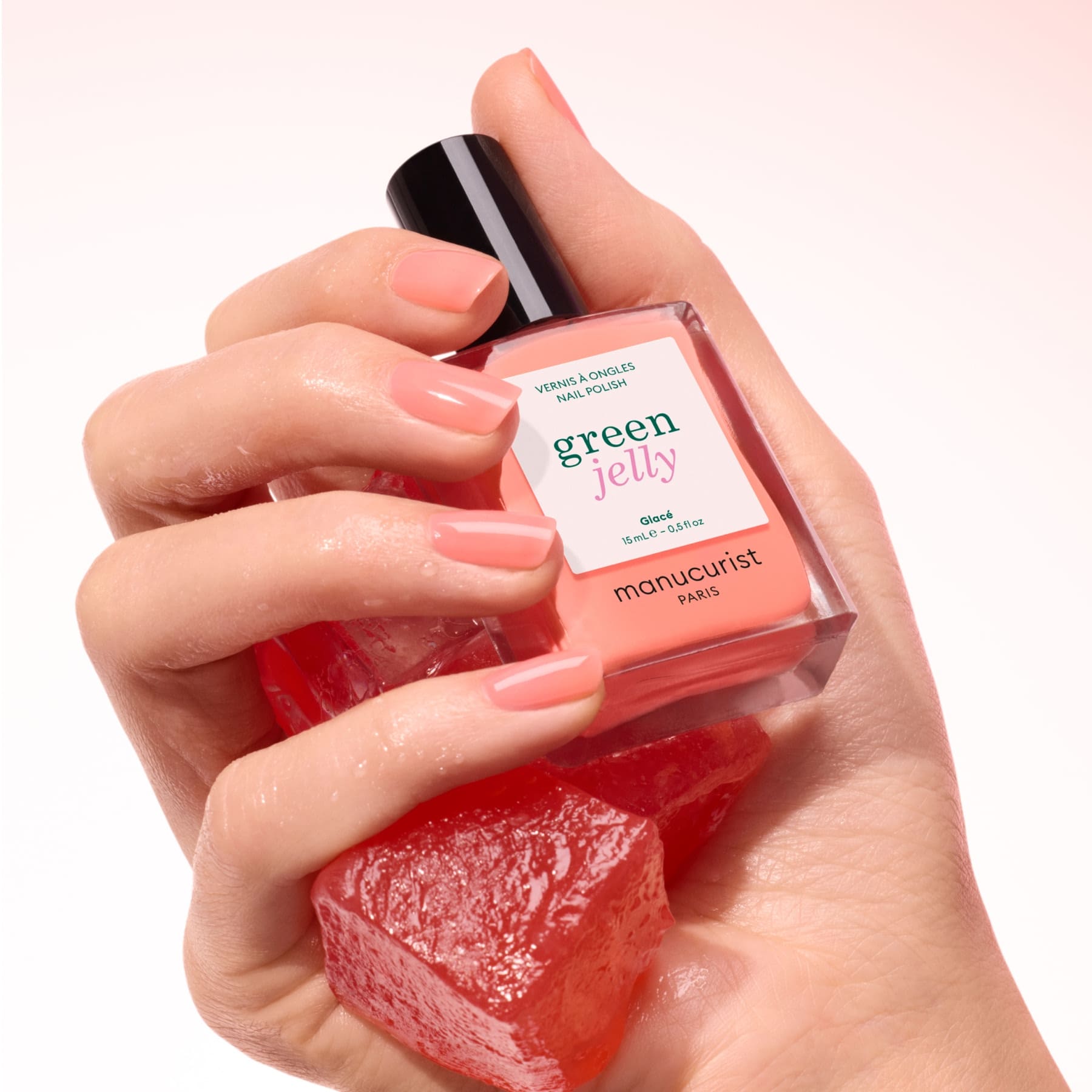A hand with neatly manicured, peach-colored nails holds a bottle of Glacé nail polish by Manucurist and a translucent red jelly cube against a soft pink background.
