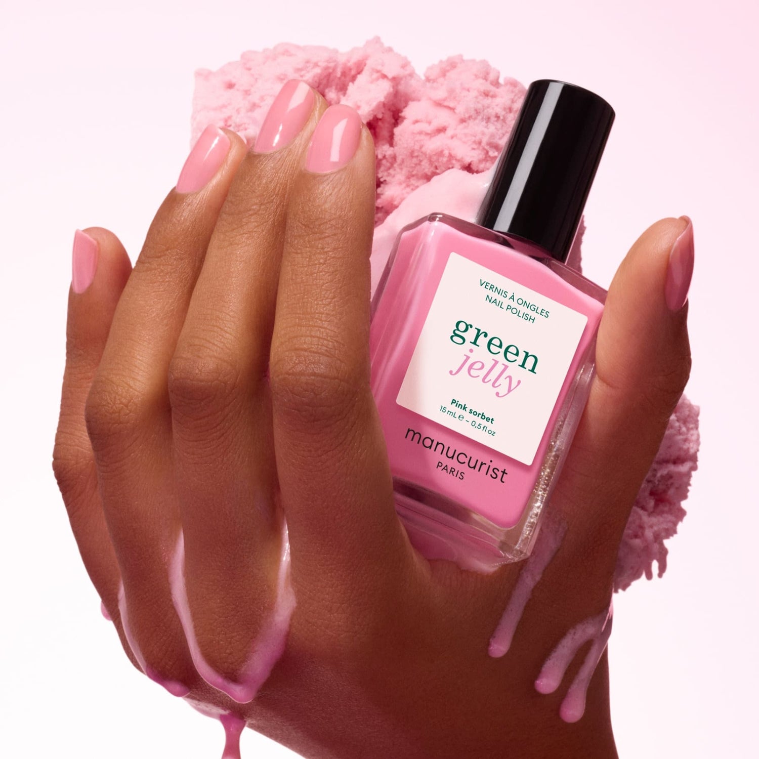 A hand with pink manicured nails displays a bottle of Manucurist Pink Sorbet Jelly nail polish, highlighting its ultra-glossy syrup effect against a soft, pink cotton-like background.