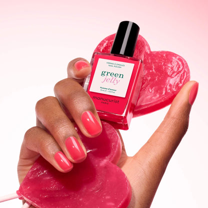 A hand with glossy raspberry pink nails holds a red heart and a bottle of Manucurist Pomme d&