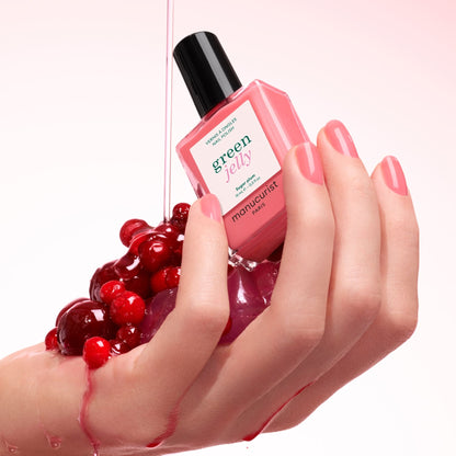 A hand with glossy, translucent pink nails holds shiny red berries and a bottle of Manucurist Sugar Plum long-lasting nail polish, as pink liquid pours over them against a soft pink background.