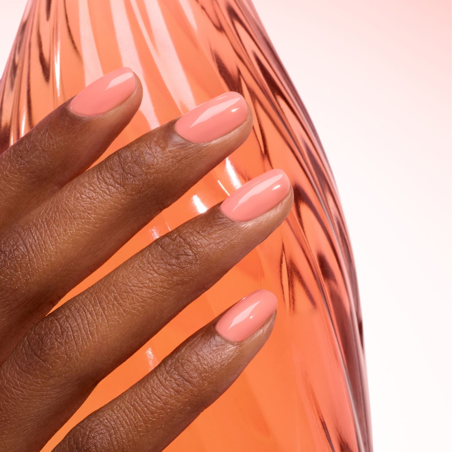 A hand with smooth, dark skin shows off neatly manicured nails painted in ultra-glossy Glacé by Manucurist, a light pink polish. The hand rests on a translucent peach-textured surface against a soft pink gradient background.