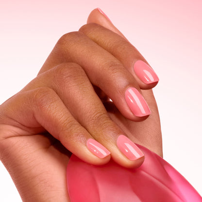 A close-up of a hand with neatly manicured nails painted in Manucurist’s Sugar Plum, a glossy translucent pink, holding a pink translucent object against a pink gradient background.