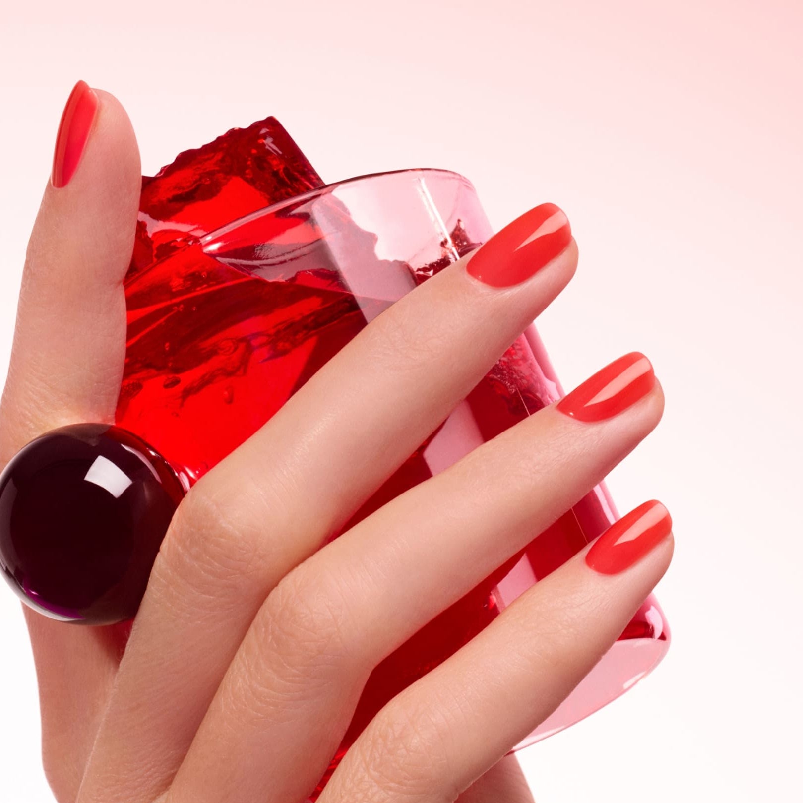 A hand with ultra-glossy Manucurist Twizzle red-orange nail polish holds a red translucent object and a pink glass, displaying a large dark purple statement ring against a soft pink gradient background.